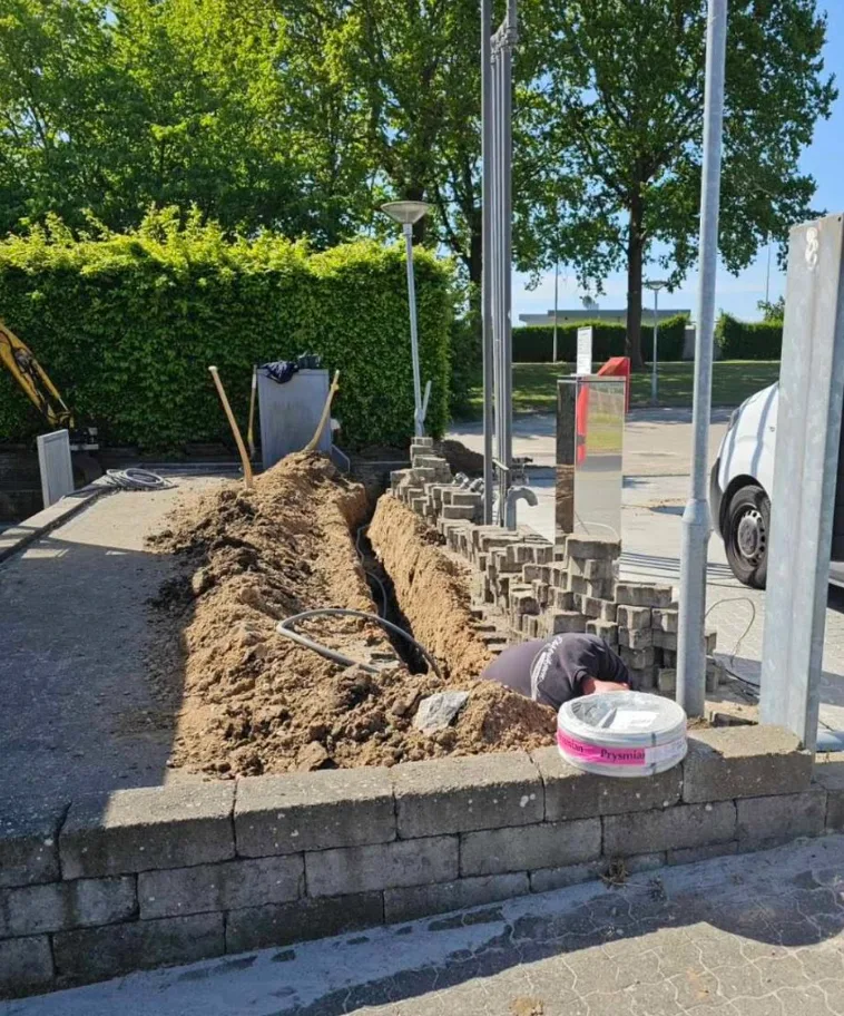 Smal kabelgrav gravet langs en belægning med opstablede fliser ved siden af
